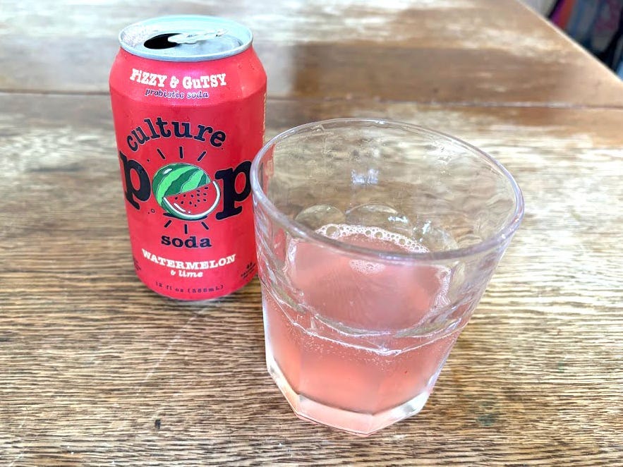 An open can of watermelon-and-lime Culture Pop next to a small, clear glass with pink liquid inside. Both are sitting on a wooden table.