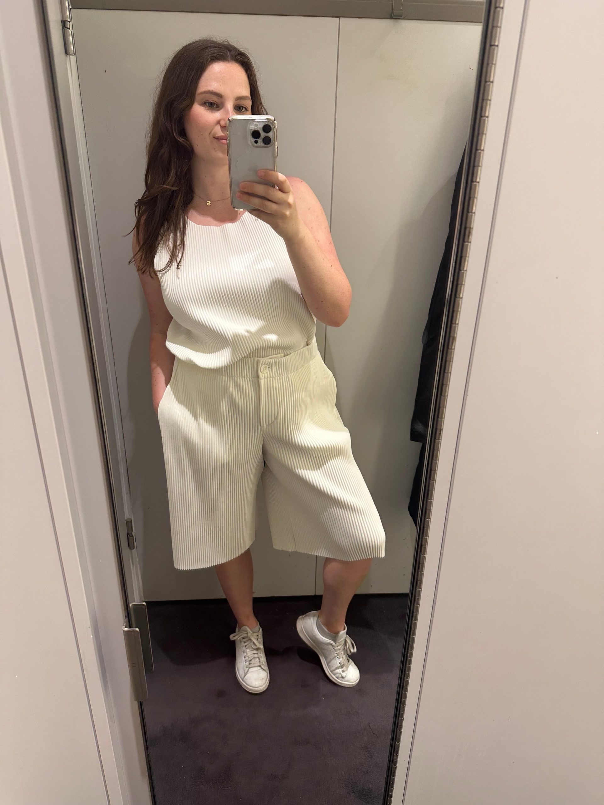 A woman takes a mirror selfie in a white top and white shorts.