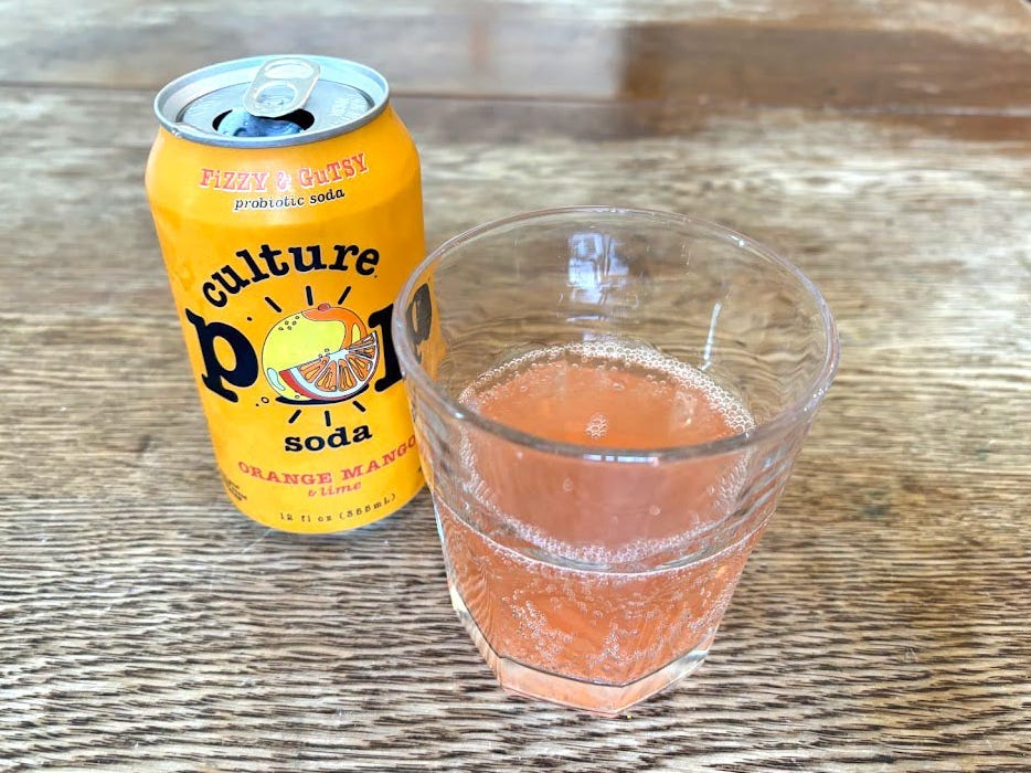 An open can of orange-mango-and-lime Culture Pop next to a small, clear glass with orange liquid inside. Both are sitting on a wooden table.