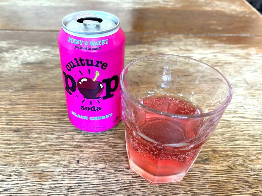 An open can of black-cherry Culture Pop next to a small, clear glass with dark-pink liquid inside. Both are sitting on a wooden table.