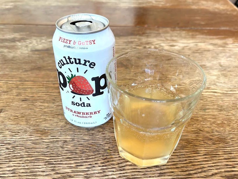 An open can of strawberry-and-rhubarb Culture Pop next to a small, clear glass with yellow liquid inside. Both are sitting on a wooden table.