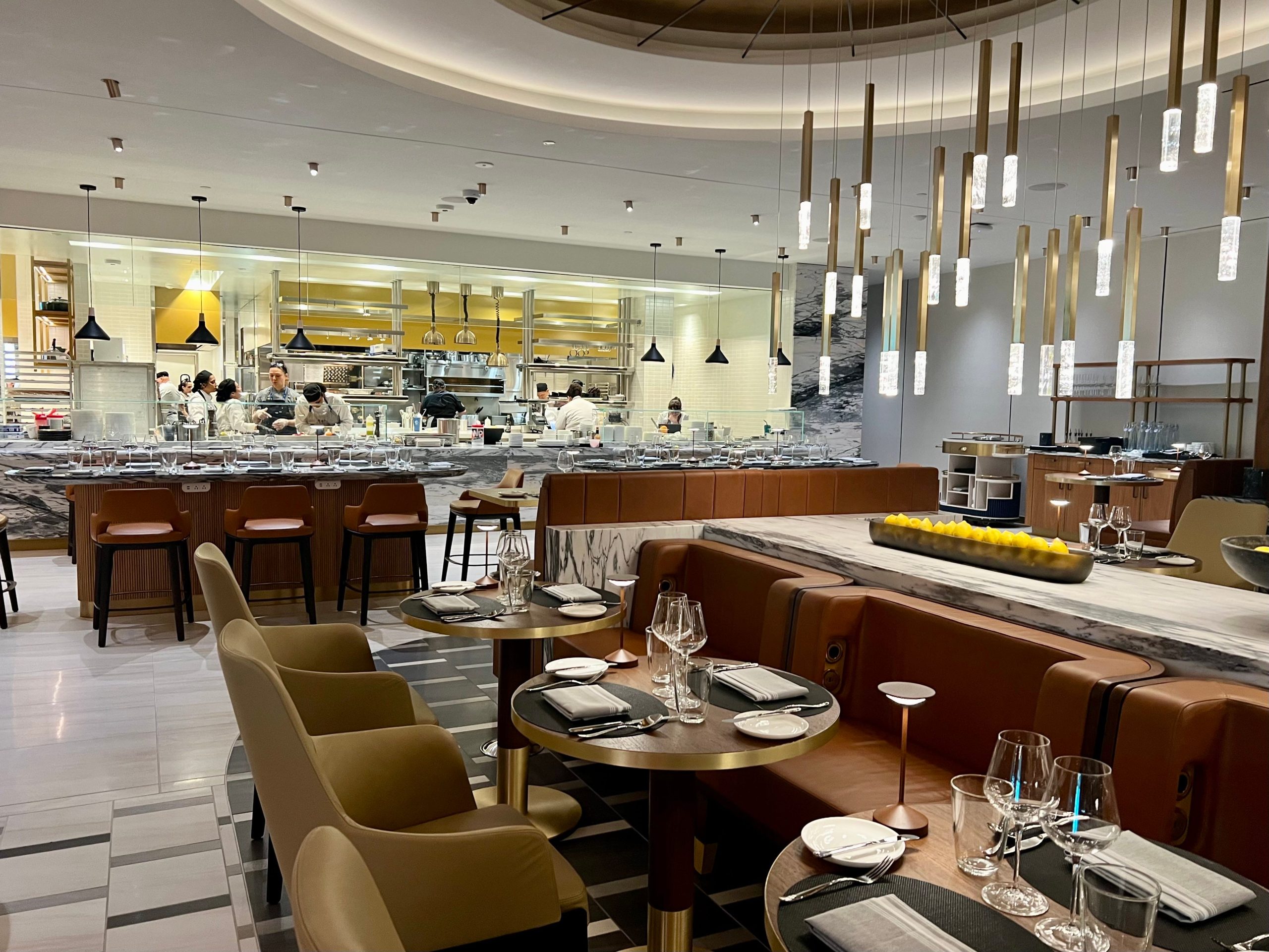 The Brasserie restaurant has beige and caramel-colored booths, chairs, and a hanging chandelier. The viewer is facing the open kitchen.