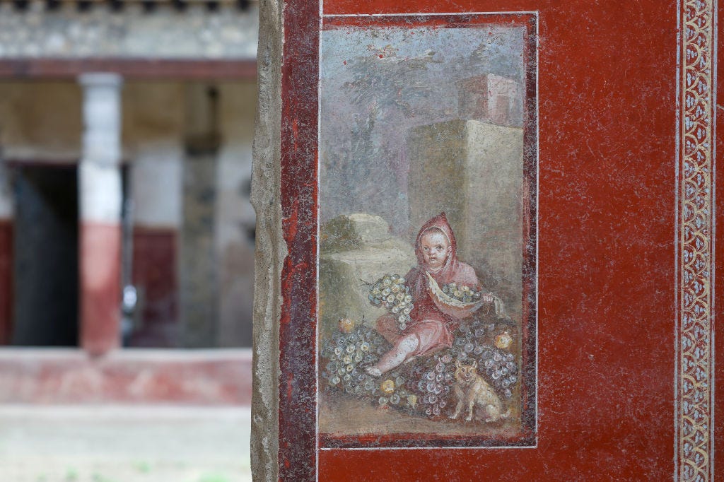 A fresco on a red wall showing a child in a red hood holding grapes next to a small dog