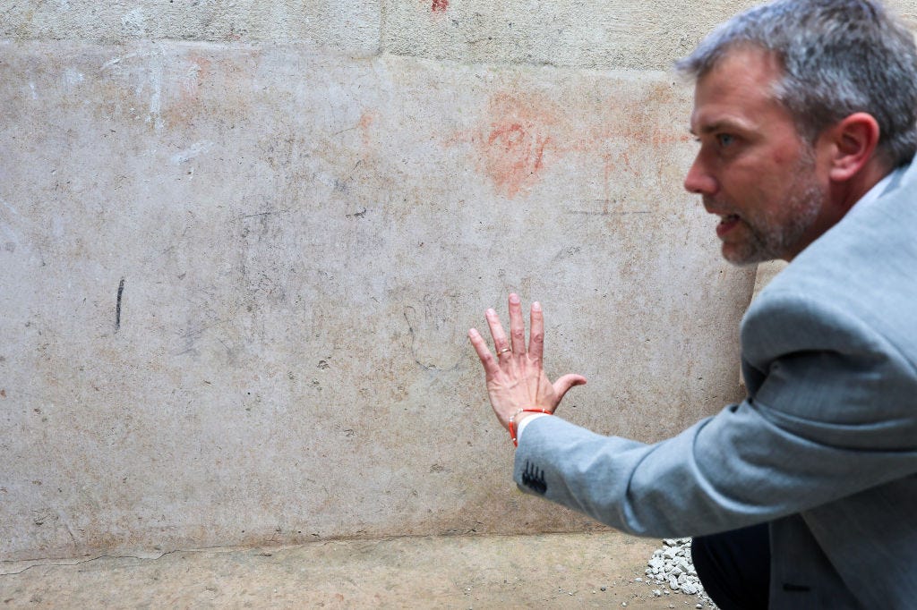 A man in a suit kneels next to a wall with a charcoal outline of a child's hand in Pompeii