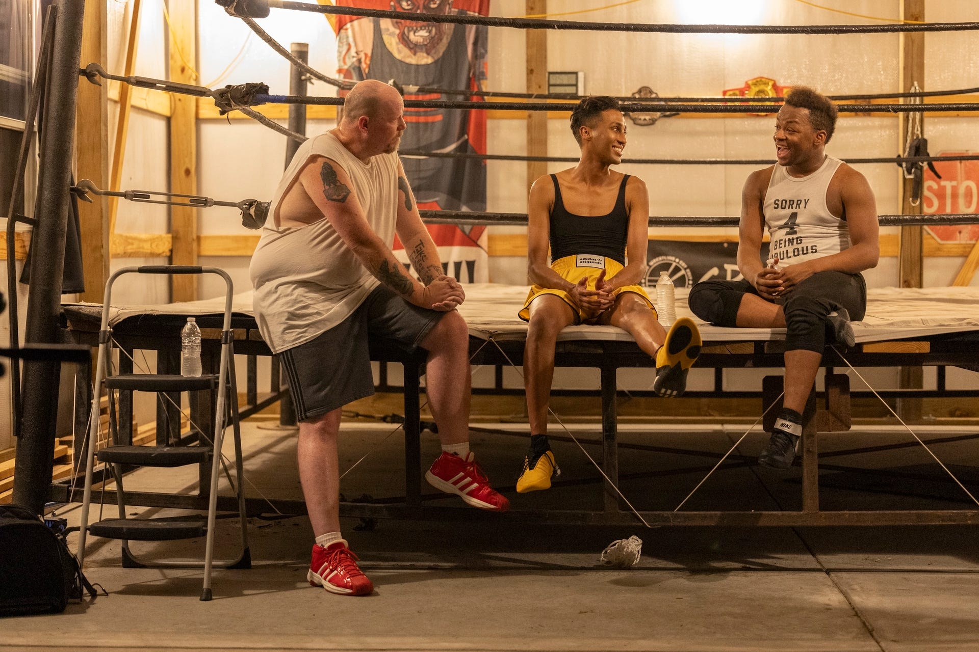 brian, priayanka, and princey on season four of we're here. they're all dressed in athletic wear, sitting casually on the edge of a wrestling while chatting and smiling