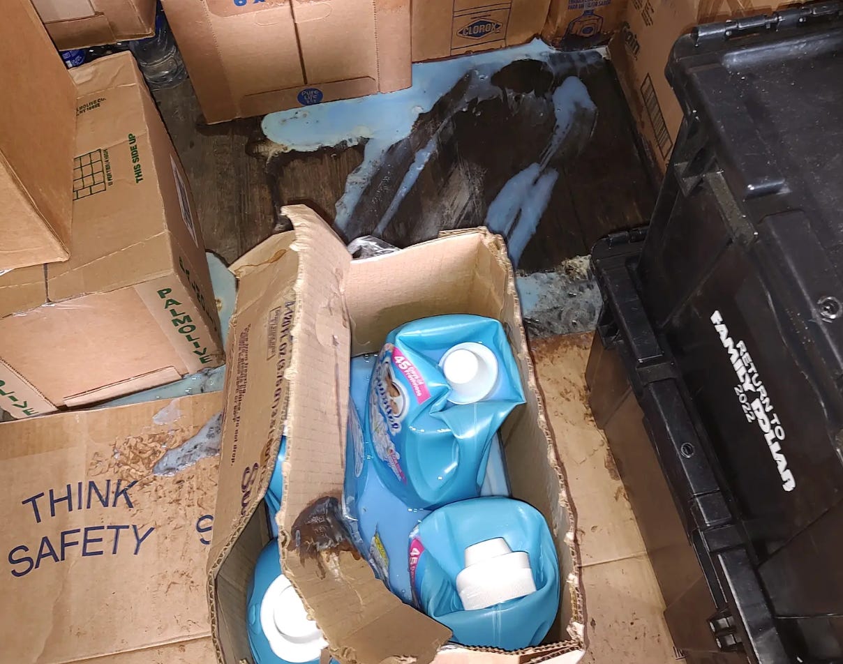 Crushed bottles of laundry detergent sit in a cardboard box with the detergent itself leaking on the floor of a trailer. A box with the caution "Think Safely" appears next to the detergent.