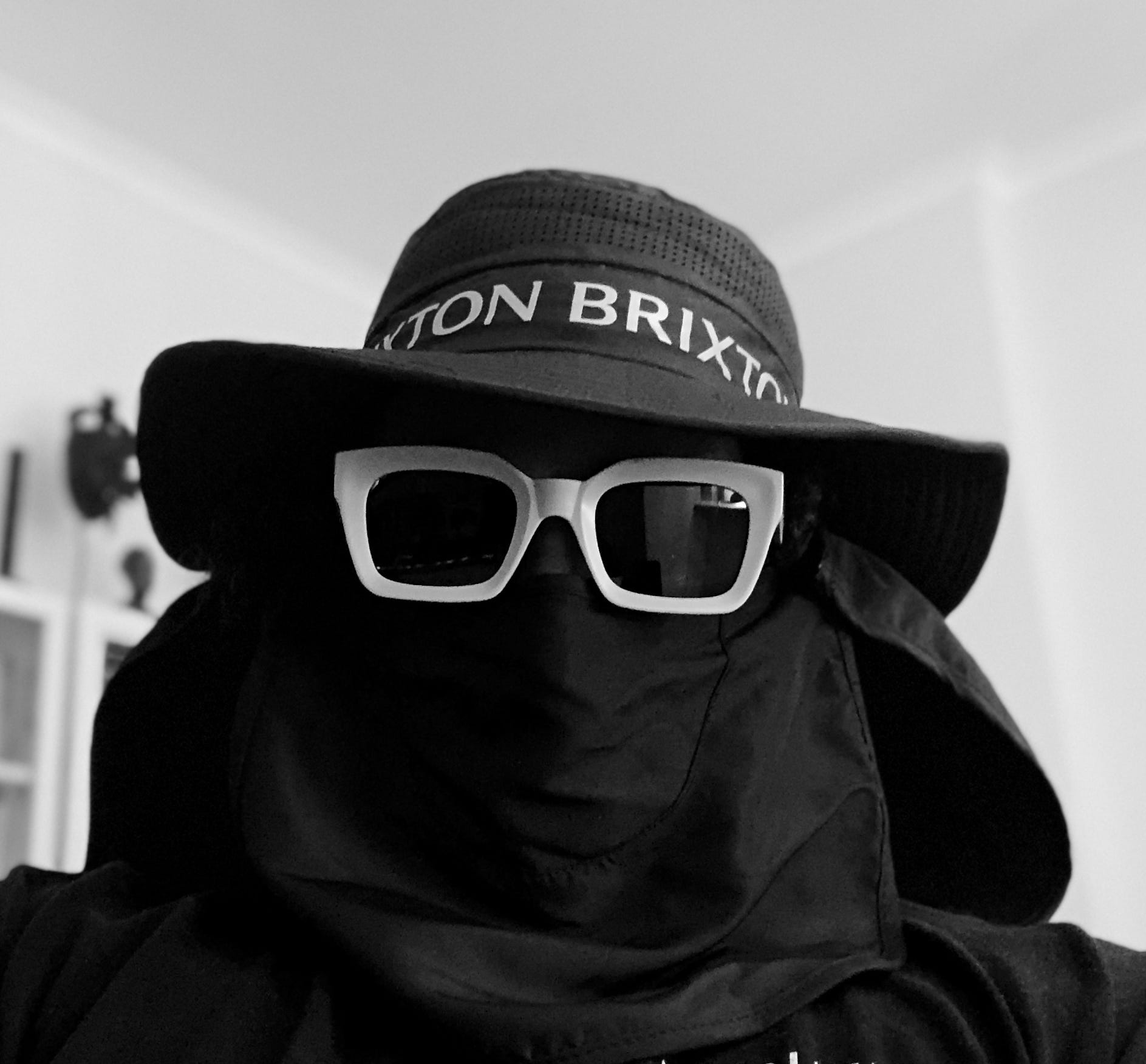 a black and white photo of the artist Vince Fraser wearing sunglasses, a wide-brim hat, and a scarf around his face