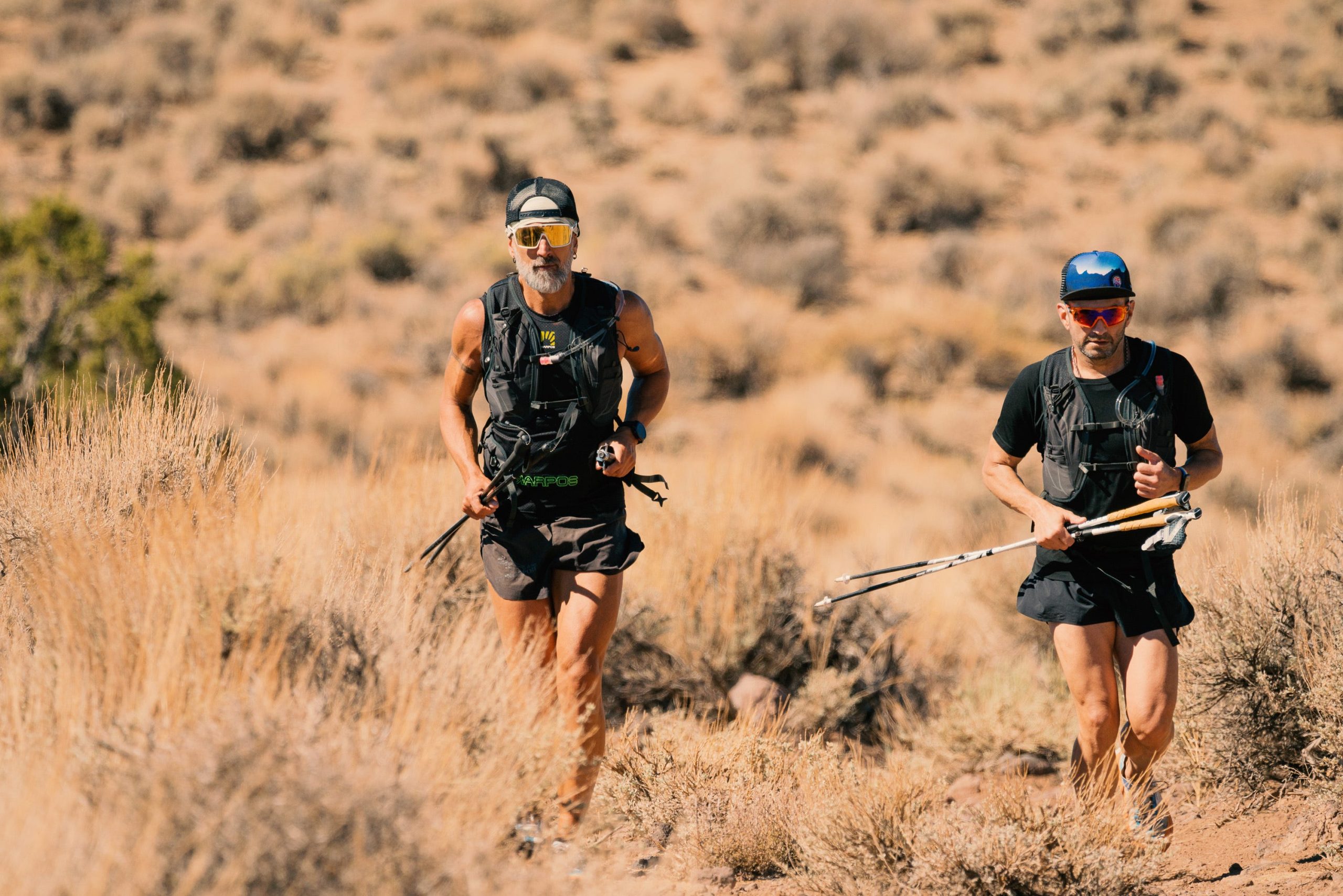 two runners in running attire with sunglasses and hats on crossing desert terrain