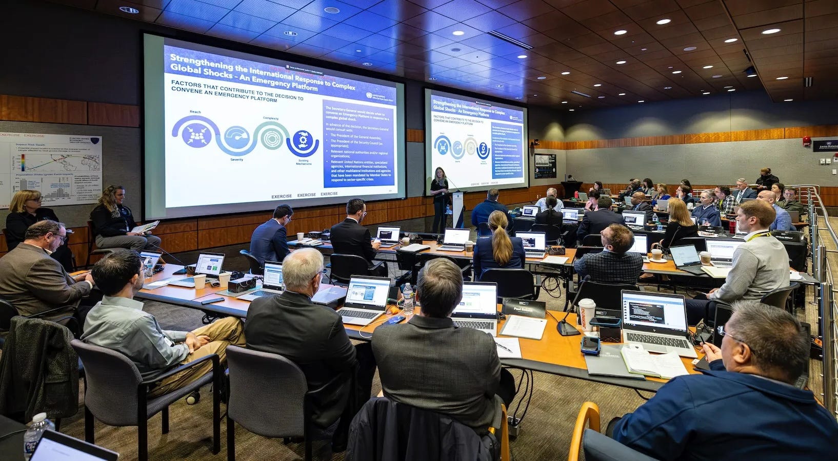 large conference room with long desks full of people in suits sitting at laptops and looking at a large presentation screen with a person at a podium presenting