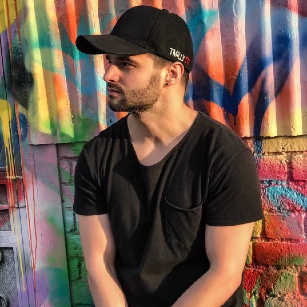 Tim Milgream wears a black T-shirt and black baseball cap standing against a rainbow graffiti wall.